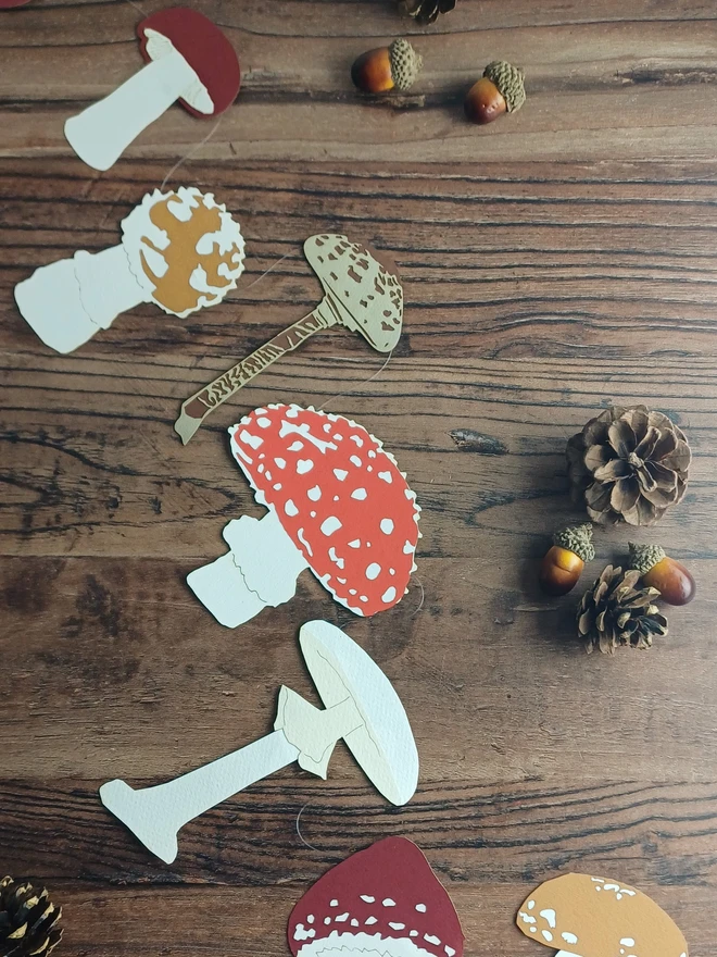 Paper Mushroom and Toadstool Garland spread on table showing 7 different designs in a mix of reds browns and whites