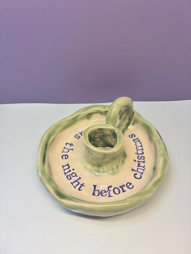 Handmade ceramic Christmas taper candlestick holder with a soft green glazed edge and the words “‘Twas the night before Christmas” in dark blue lettering, arranged around the candle holder opening. Photographed against a pale background.