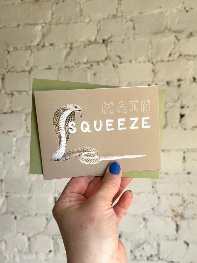 a beige greetings card featuring an illustration of a cobra with the text “main squeeze” with a green envelope