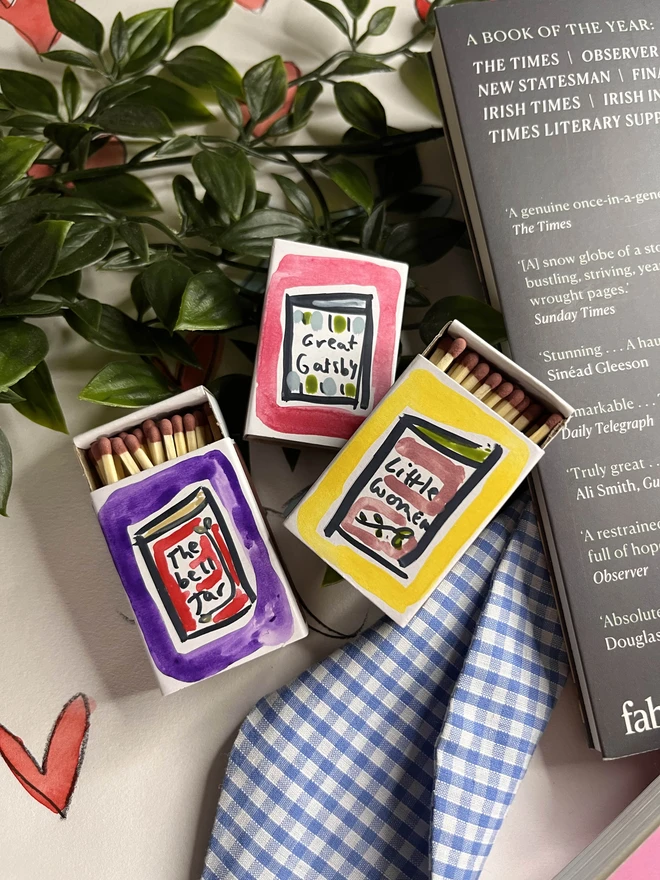 A collection of three hand-painted matchboxes with playful and colorful designs: one features two glasses with the word "Cheers!" written in bold lettering, another displays bright yellow lemons with "Easy Squeezy" written alongside, and the third showcases a red bow with the phrase "Bows & Arrows." Each matchbox is crafted with vibrant illustrations and creative lettering.