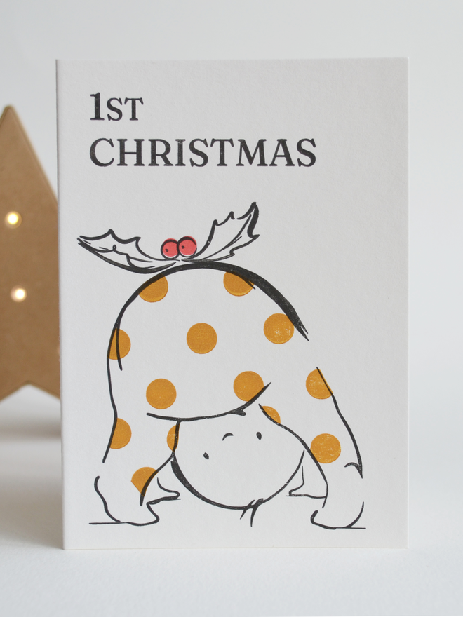 Baby's first christmas greeting card with a cute baby playing peekaboo through legs in a spotty outfit with a holly sprig on top to resemble a christmas pudding.