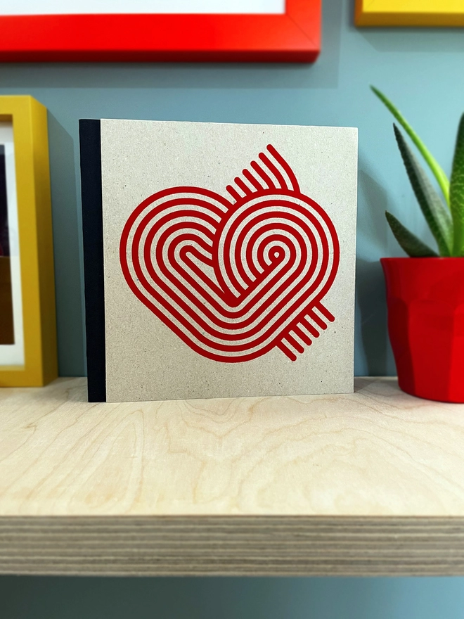 Stripy Heart design is screenprinted in magenta on a grey pasteboard sketchbook with a black fabric spine, stood on a plywood shelf with hints of framed pictures around. the wall is duck egg blue, a plant glimpsed to the side.