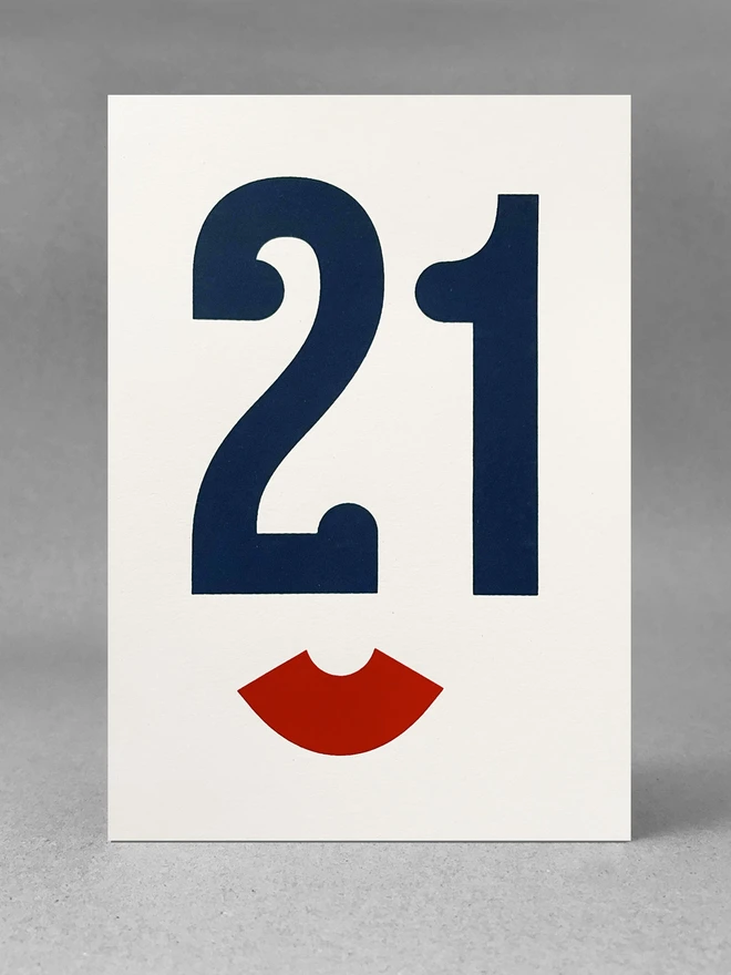 The Number 21 screen printed in a dark blue serif typeface. The serifs make the number 21 look like the eyes and nose of a face from a slight angle. Below is a red bracket on its side. This looks like the lips of the face. The image has an Art Deco feel to it. The portrait card is stood straight on in a light grey studio set.