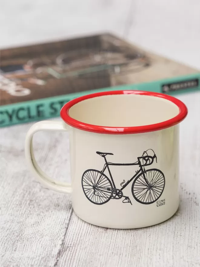 Picture of a Cream Enamel Mug with a Red Rim with a Bicycle design etched onto it, taken from an original Lino Print