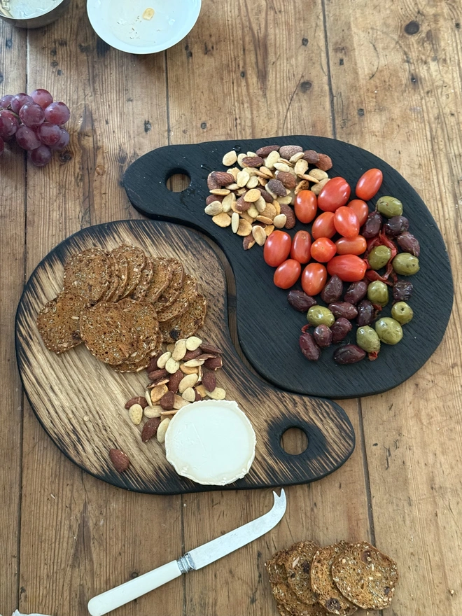 “Pair of handmade Yaki Sugi serving boards in blackened and natural oak, styled together with olives, cherry tomatoes, grapes, crackers and cheese on a rustic wooden table.”
