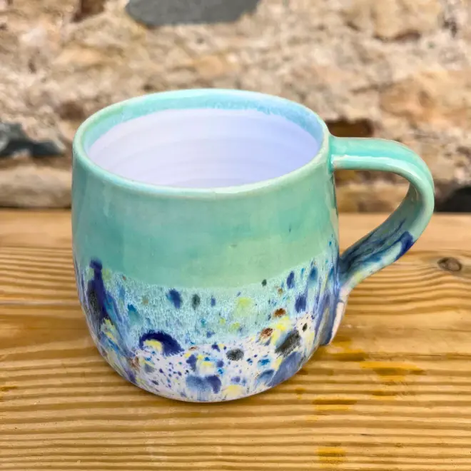 Shoreline Large Round Mug, a colourful mug on a wooden surface against a brick backdrop. 