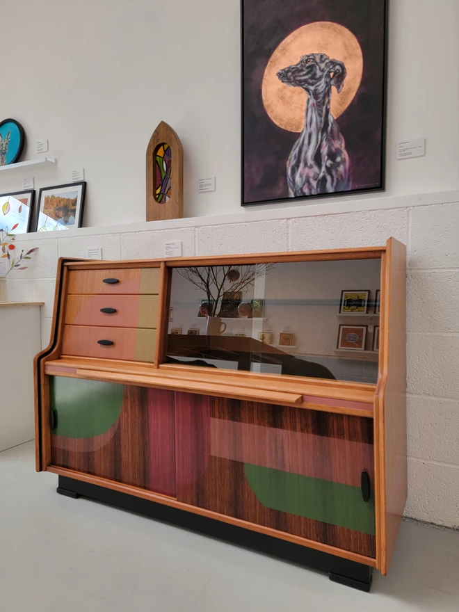 Upcycled mid-century teak sideboard with pink and green shapes and glass doors on display in a gallery surrounded by art
