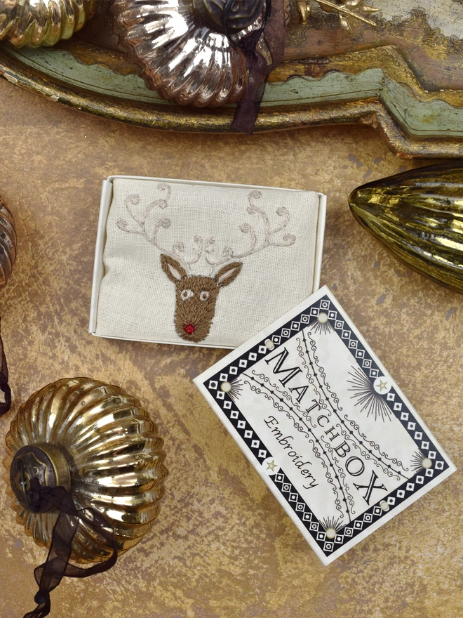 Rudolph the red nose reindeer with a red crystal nose, inside a Matchbox displayed on a gold background with glass baubles.