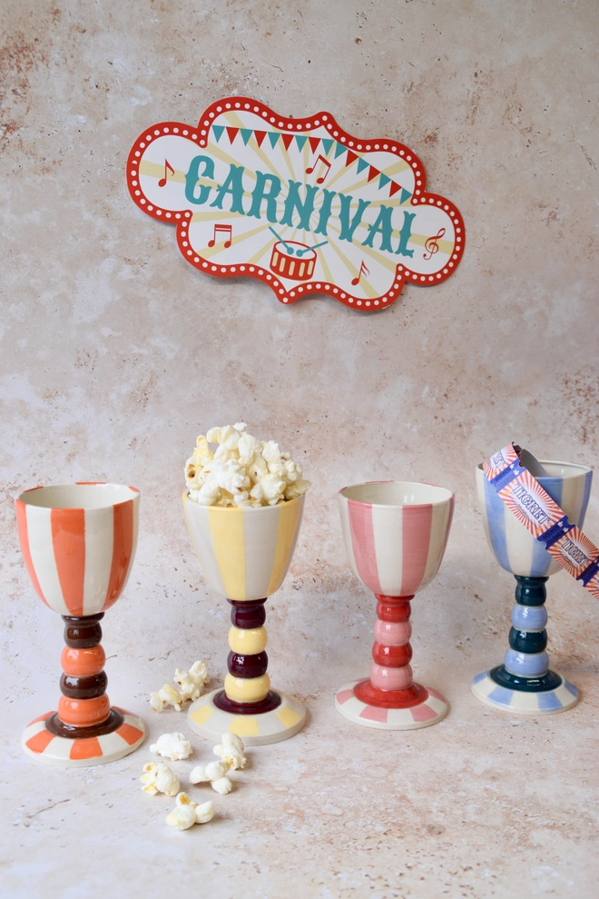 Handmade striped ceramic goblets in orange, yellow, pink and blue colourways, styled with popcorn and sweets beneath a “Carnival” sign on a neutral background.