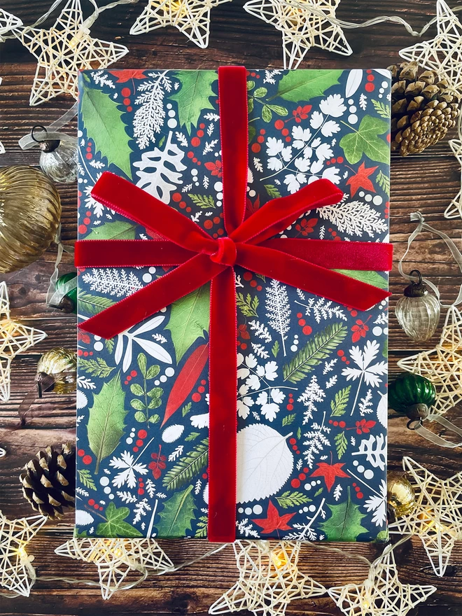 Christmas present wrapped in recycled and recyclable gift wrap with Winter foliage design, red ribbon, surrounded by star lights and baubles on wood background