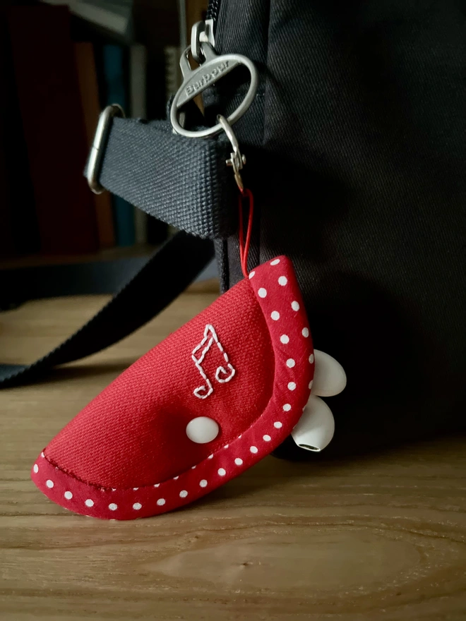 Soft cotton red earphone case with polka dot trim and white snap closure, embroidered with musical notes, holding earphones and attached to bag.