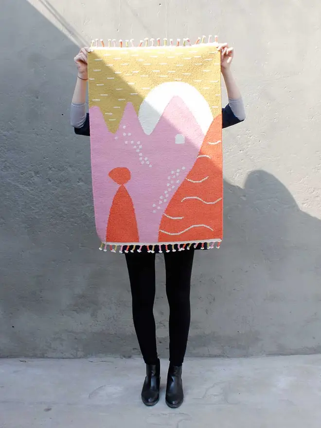 Landscape Flatweave Rug, a person holding a colourful rug against a grey stone backdrop. 