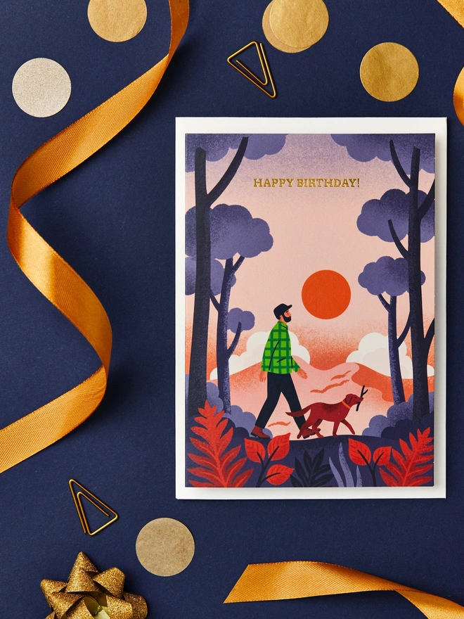 Greeting card with illustration of a bearded man walking in a forest with a dog. The card is on a desk with a navy blue background and gold ribbon decorations.