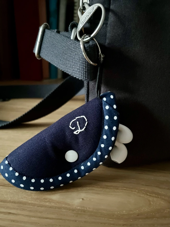 Soft cotton navy earphone case embroidered with musical notes, with polka dot trim and snap closure, holding white earphones, attached to bag.