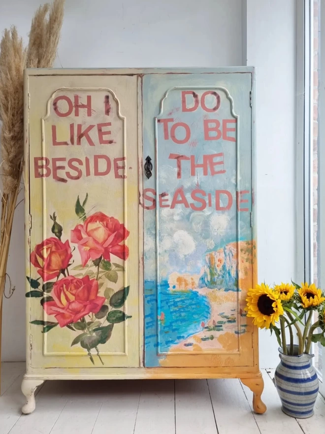 Upcycled painted cupboard or gentleman’s wardrobe. Featuring a seascape Monet painting and a vintage rose image in the shabby chic style. One door is cream, one is blue and wording reads in pink ‘Oh I do like to be beside the seaside’.