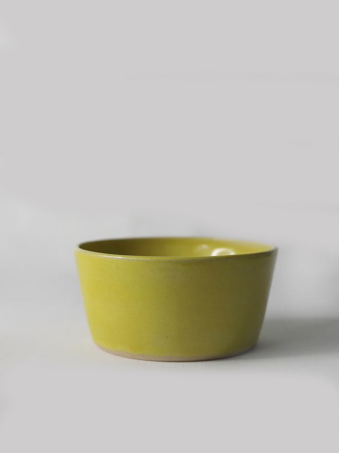 A single hand-thrown ceramic breakfast bowl in sun yellow, with a glossy glaze showing subtle variation in tone, a gently flared form and unglazed natural clay base, photographed against a plain light grey background.