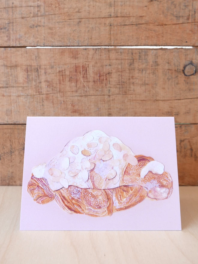 The front of the Almond Croissant Card depicting an almond croissant on a pale pink background 