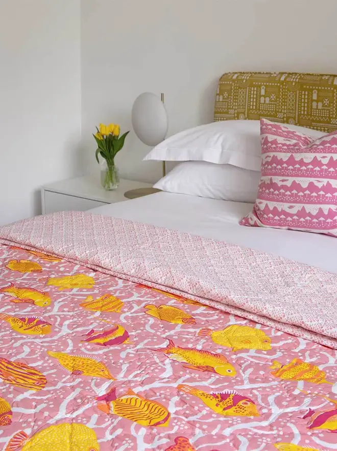 Pink Coral Reef Quilt, a pink quilt sitting on a bed. It is accompanied by other bedroom items such as a vase and some pillows. 