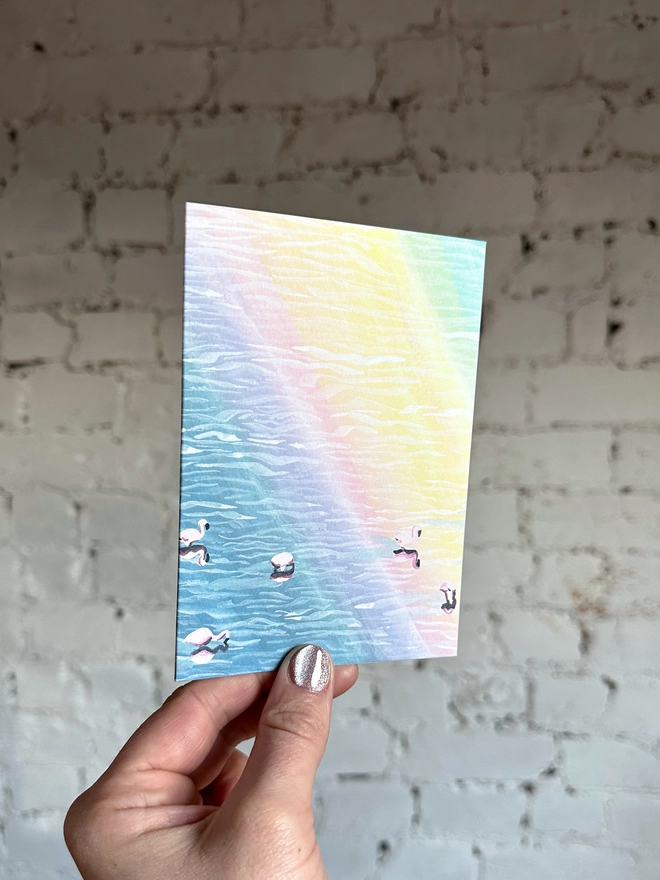 a greetings card featuring an illustration of flamingos in water with a rainbow reflection