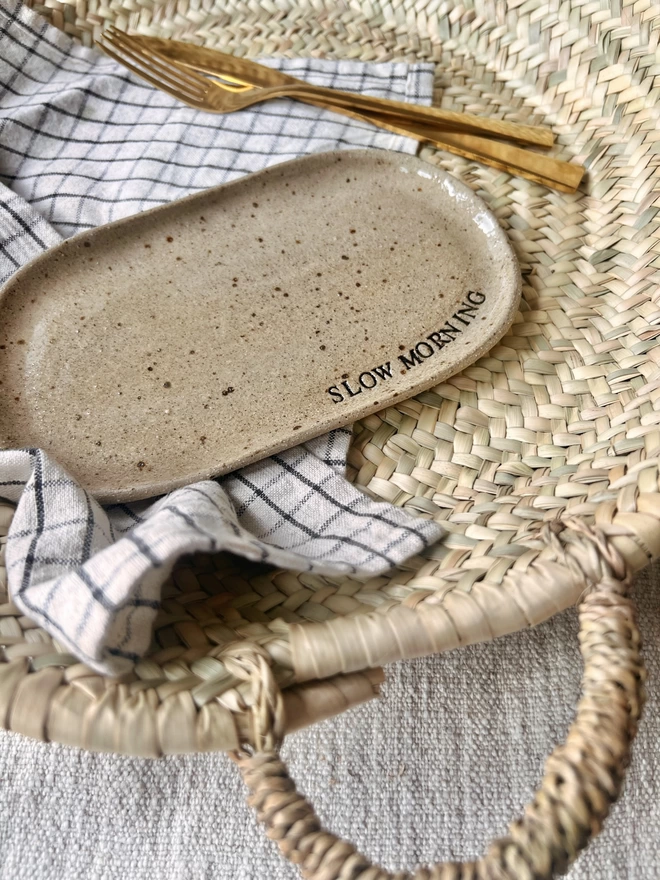 oval ceramic plate laid on a chequered tea towel beside a gold knife and fork. Plate has the words 'slow morning' stamped in black around the edge