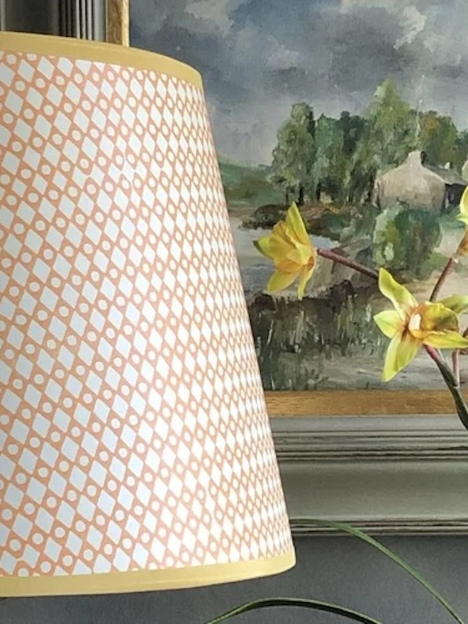 Retro Drum Lampshade. Close-up of a peach and white diamond lattice lampshade with a soft yellow trim beside daffodils and a landscape painting. The hand-printed shade adds a fresh, springlike touch to a mantelpiece or side table.