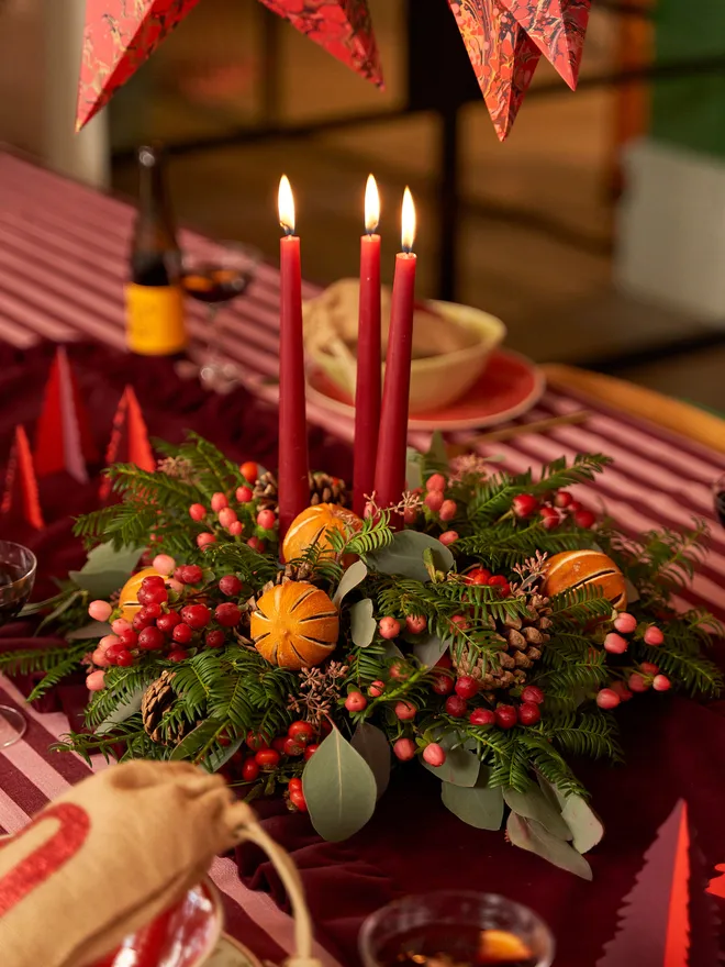 green wreath with dried oranges, red berries, and leaves on a table with red striped table cloth. three lit, red candles in the middle