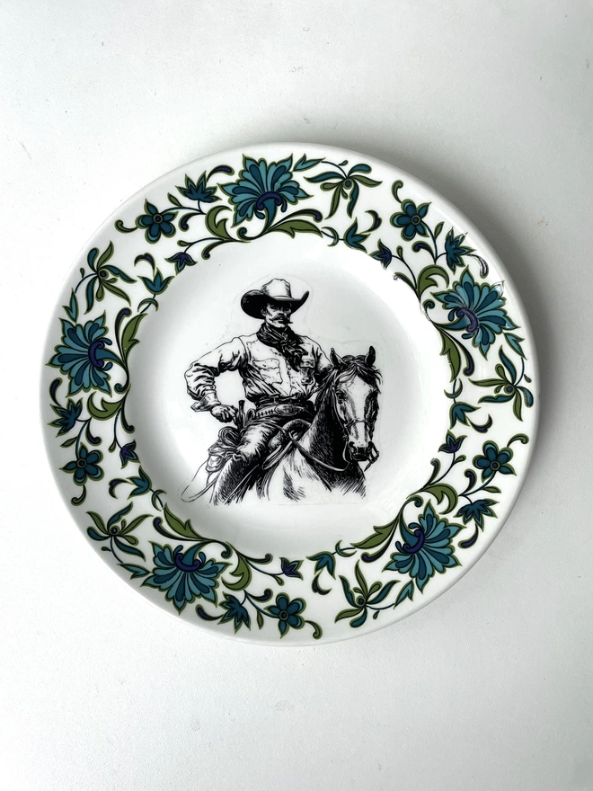 vintage plate hanging on a white wall, it has a decorative border, with a printed vintage illustration of a cowboy in the middle 