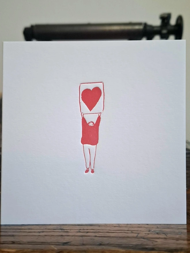 Heart Letterpress Handprinted Greetings Card, a white heart card with a red design sitting on a wooden surface. 