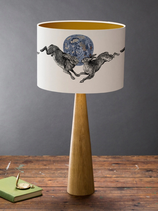 Drum Lampshade featuring a pair of hares leaping across a blue and silver moon on a wooden base on a shelf with books and ornaments