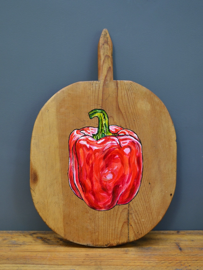 Wooden chopping board with handpainted design of a red pepper standing against a wall