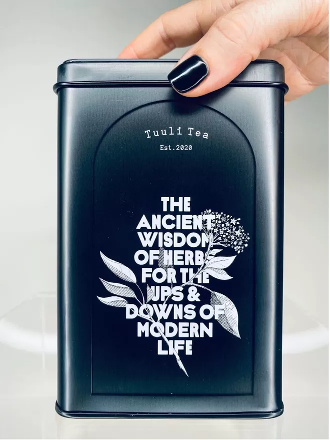 A black Tuuli Tea tin with white writing saying the ancient wisdom of herbs for the ups and downs of modern life. There is a hand with black nail polish holding the top of the tin. 