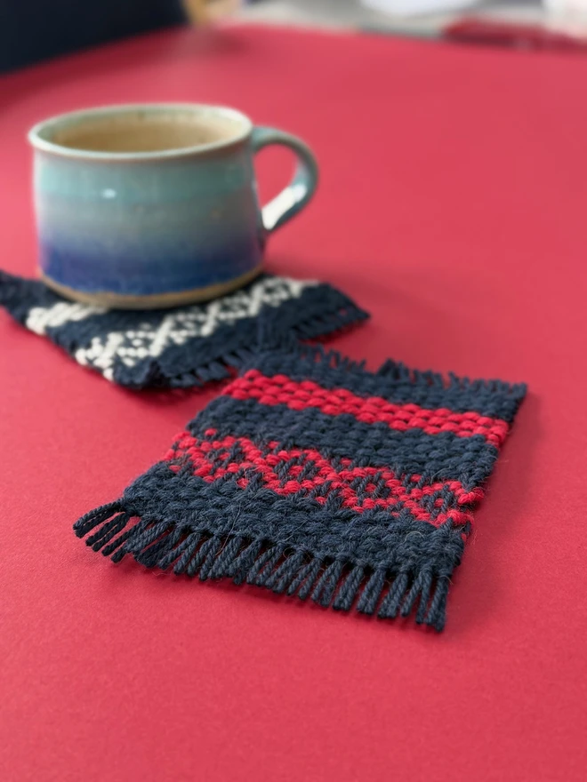 Two navy, red and cream wool coasters with a blue mug on a red background