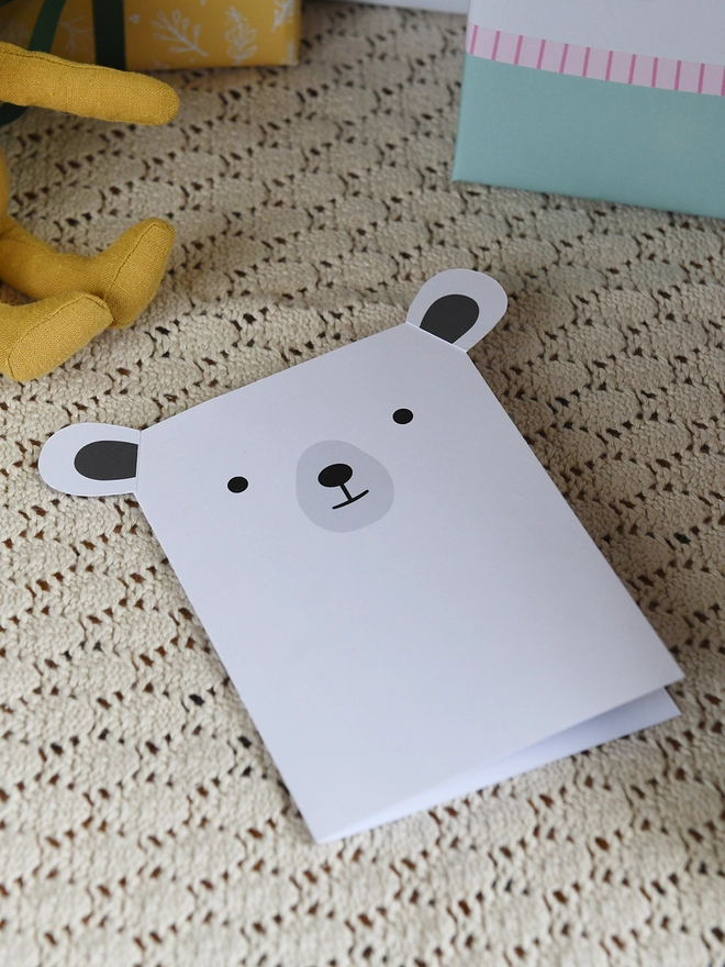 A white polar bear greetings card with stick on ears lays on a cream blanket.