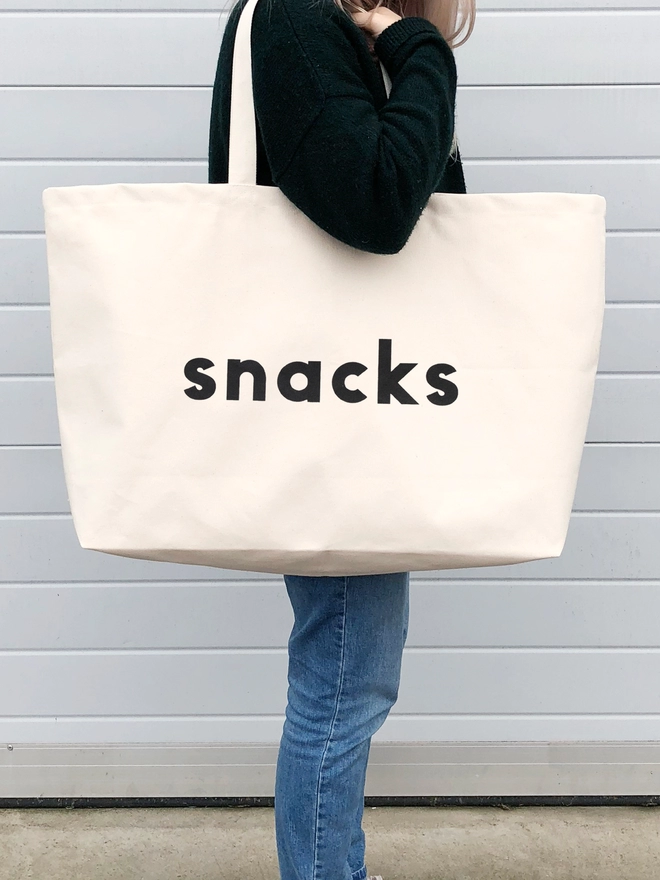 model carrying an oversized natural canvas tote bag with bold snacks slogan