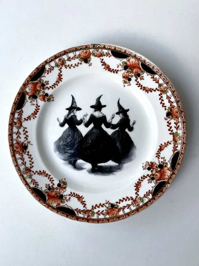 a coven of 3 witches dance hand in hand, the image is victorian in black and white and is printed directly onto an ornate vintage plate