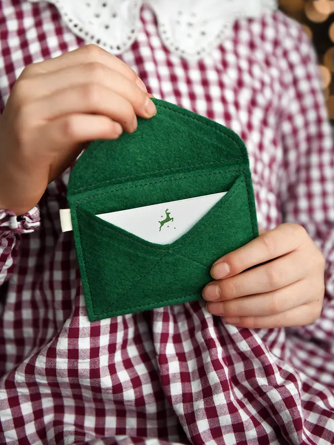A child wearing a red gingham dress is holding a handmade green felt envelope with a festive notecard tucked inside.