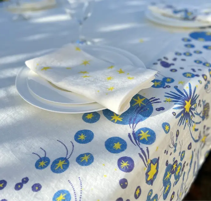 Solaris Tablecloth In White Or Blue. White tablecloth with blue and yellow starburst motifs, styled with white plates and a matching napkin.