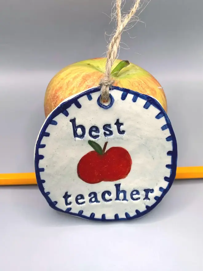'Best Teacher' Handcrafted Ceramic Ornament, a ceramic apple ornament sitting on a plain white surface joined by an apple. 