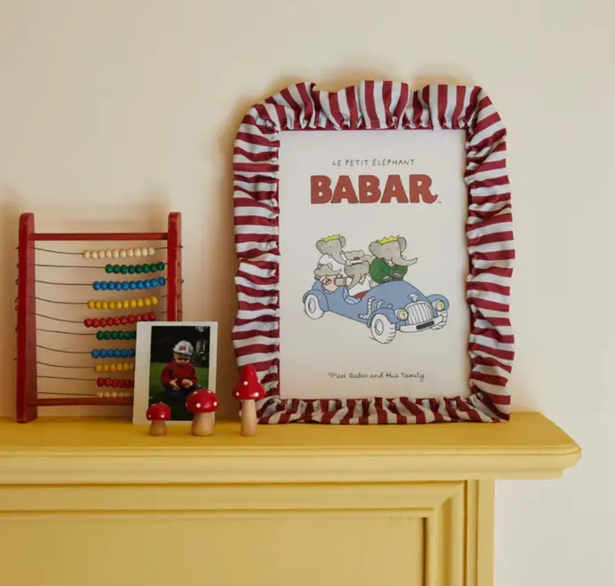 Soft ruffle frame with red and white striped fabric trim displaying a Babar print on a yellow dresser beside an abacus and small ornaments.
