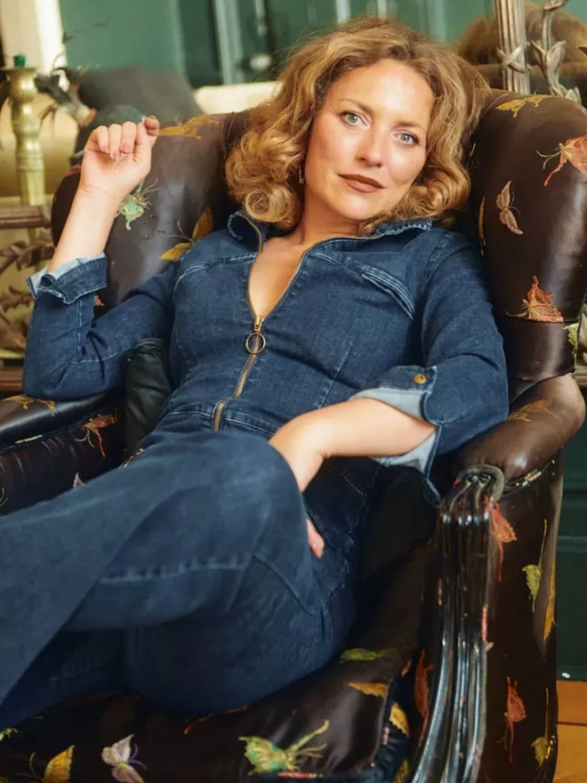 model sitting on arm chair wearing the ruby denim jumpsuit with long sleeves