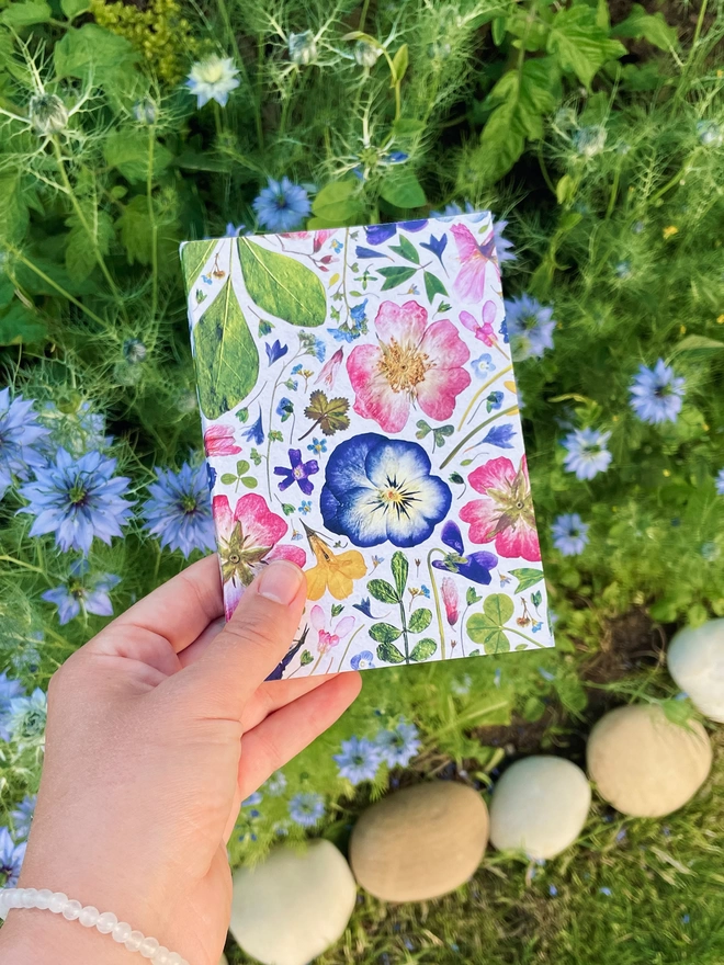 Hand Holding A6 Notebook with Pressed Flowers Design on Garden Background. Cover with Pressed Hydrangea, Viola, Cornflower, Lavender, Dog-Rose, Daisy, Verbena, Buttercup, and Forget-Me-Not