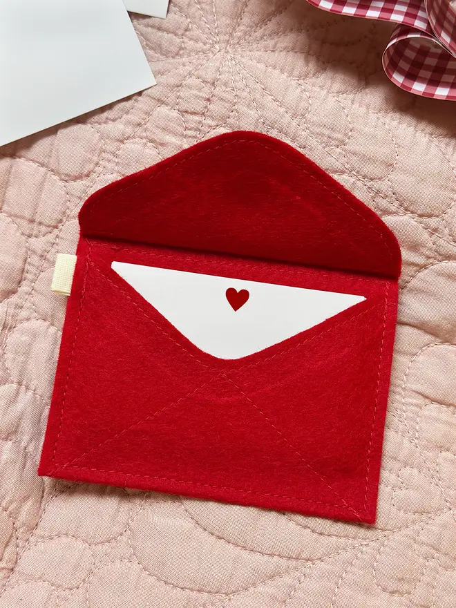 A red felt envelope is lying face down on a pink quilt, with a small stack of heart cards tucked inside.