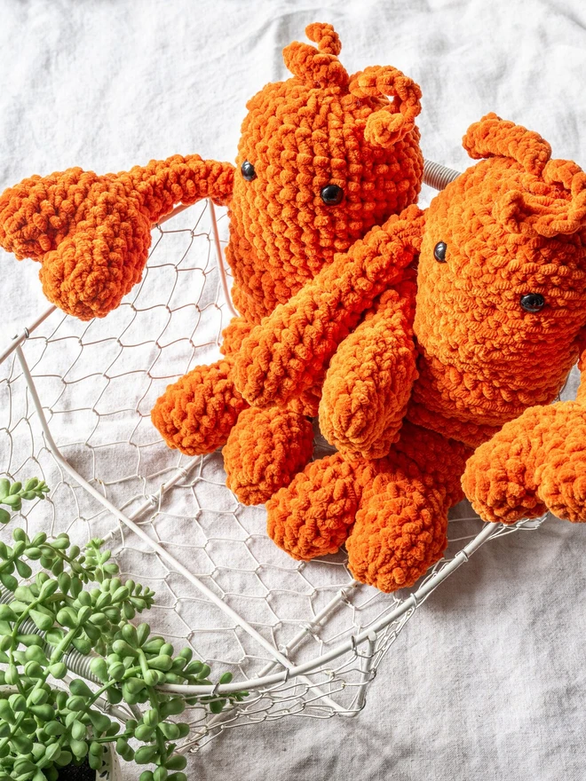 Chunky Chenille Cuddly Crochet Crustacean Lobster Plush, an orange lobster plush sitting next to another lobster on a white surface accompanied by a bag and some greenery. 