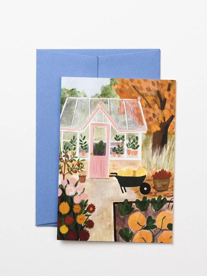 A greetings card featuring an autumnal garden. There is a glasshouse and tree in full autumn leaf in the background. In the foreground there is a dahlia patch and a pumpkin patch.