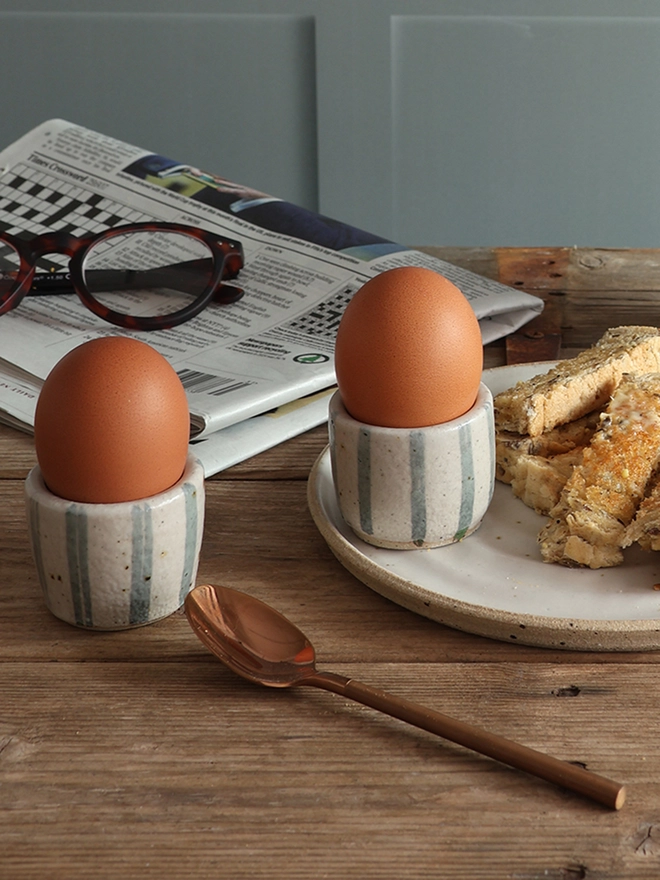 A breakfast scene featuring two handmade ceramic egg cups with a blue and white stripe design holding boiled eggs. One of the egg cups sits on a white handmade ceramic plate filled with buttered toast. A brass spoon and a newspaper with a pair of glasses on top also sit on the table.  