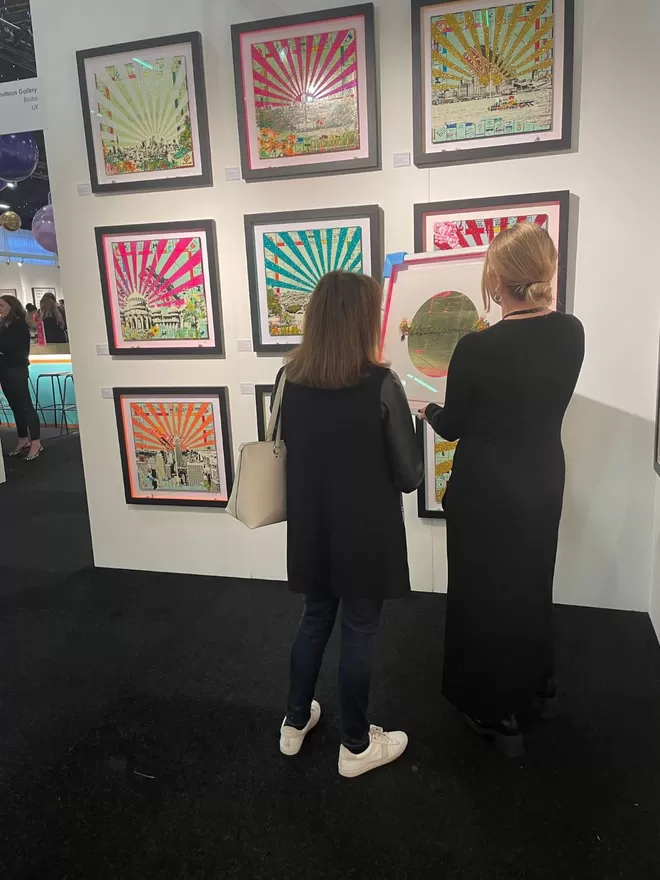 Two ladies in a gallery standing in front of a wall of framed Monopoly board prints 