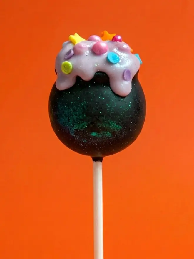 Black Witches Cauldron cake pop with colourful sprinkles on a white stick against a bright orange background