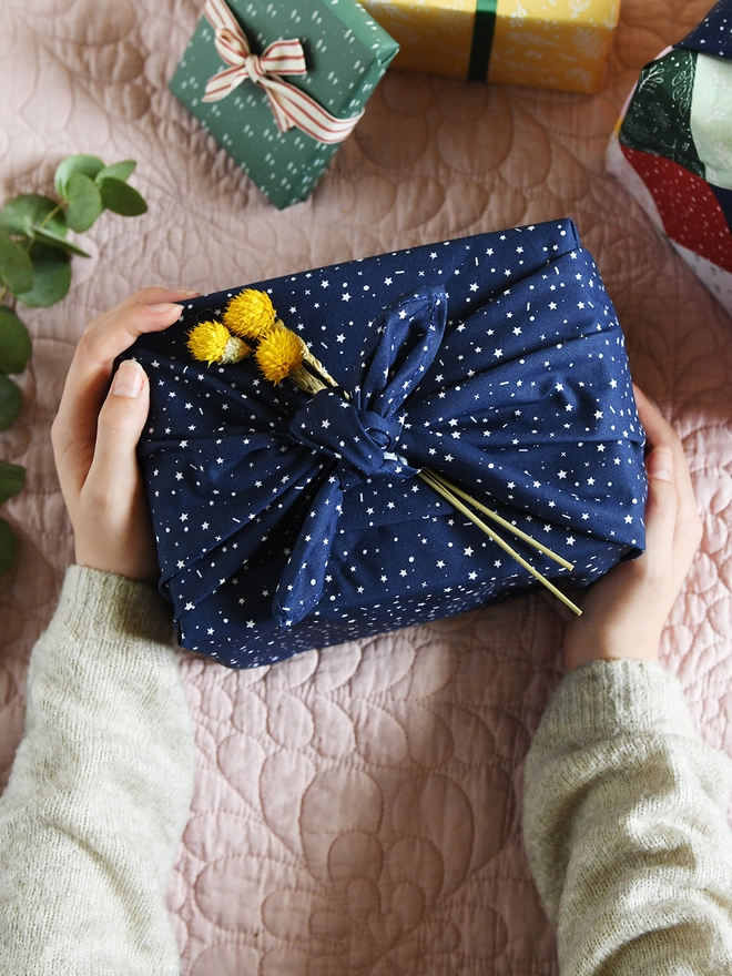 A gift is being wrapped in navy blue cotton fabric wrap with a white star design, and a small posy of yellow flowers is tucked into the knot.