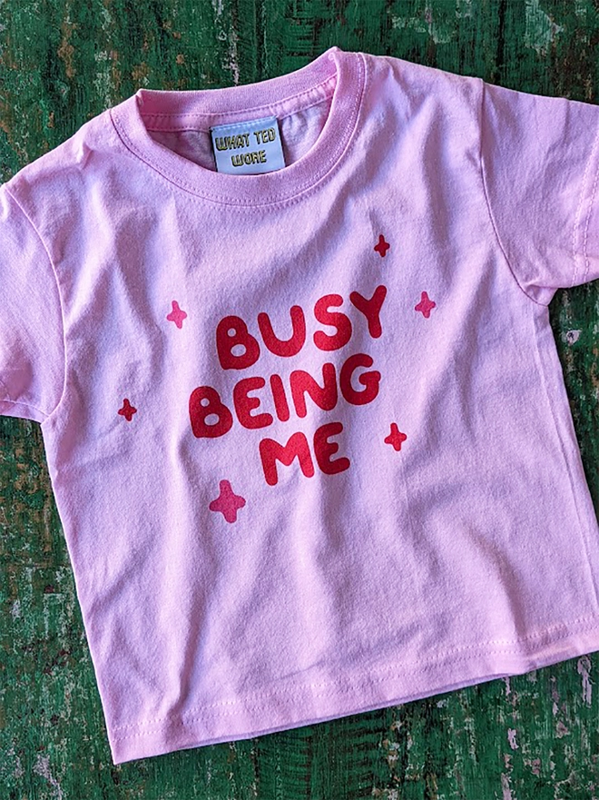A pink t-shirt with red Busy Being Me slogan on a green background