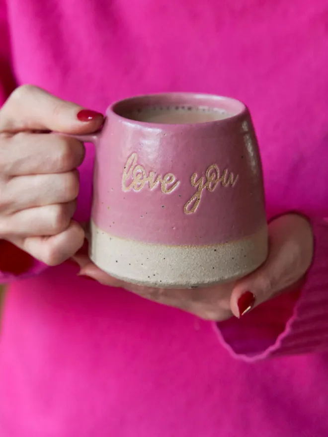 Love you ceramic pink mug. A pink mug with love you written on the front held by someone wearing a pink jumper.